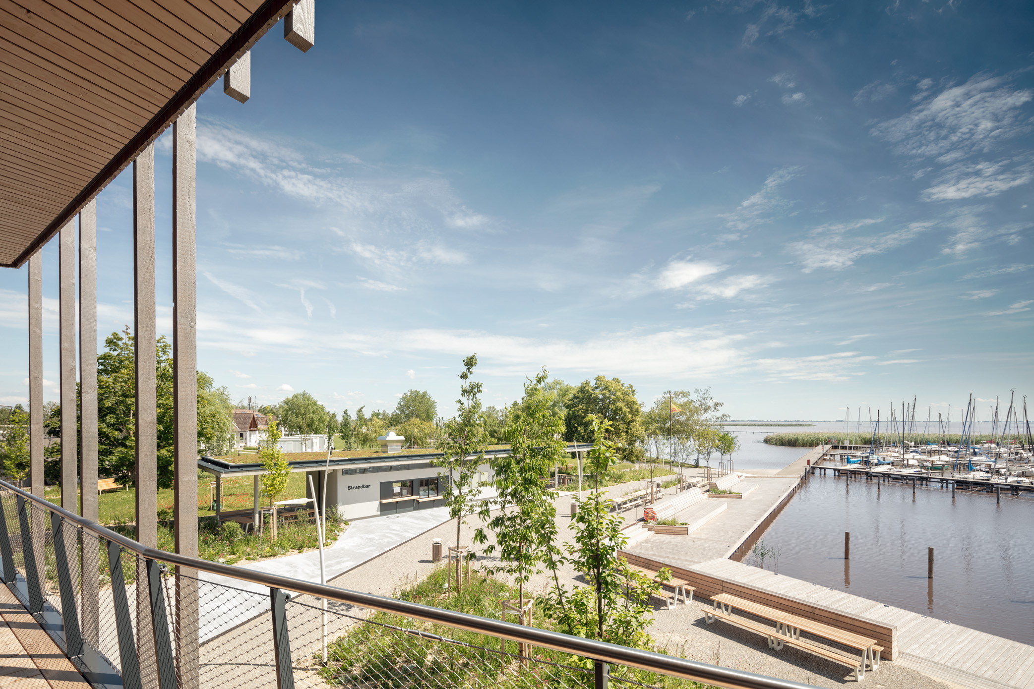 Weitläufiger Blick von einer modernen Holzterrasse auf eine Uferpromenade mit Sitzbänken, Bäumen und einem kleinen Hafen mit Segelbooten – unter klarem Himmel und ruhiger Atmosphäre.