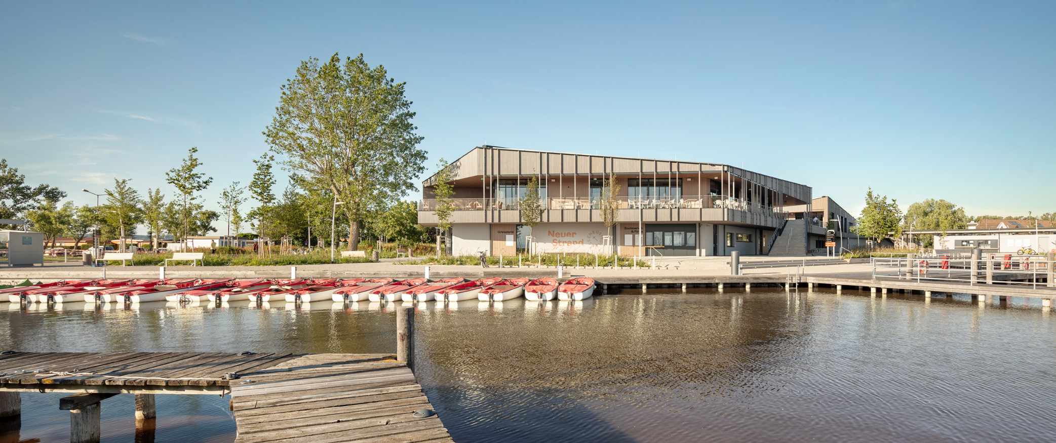 Modernes Hafengebäude Neuer Strand mit Holzfassade am Ufer, umgeben von Bäumen und roten Ruderbooten, unter klarem blauen Himmel – ideal für Freizeit und Wassersport.