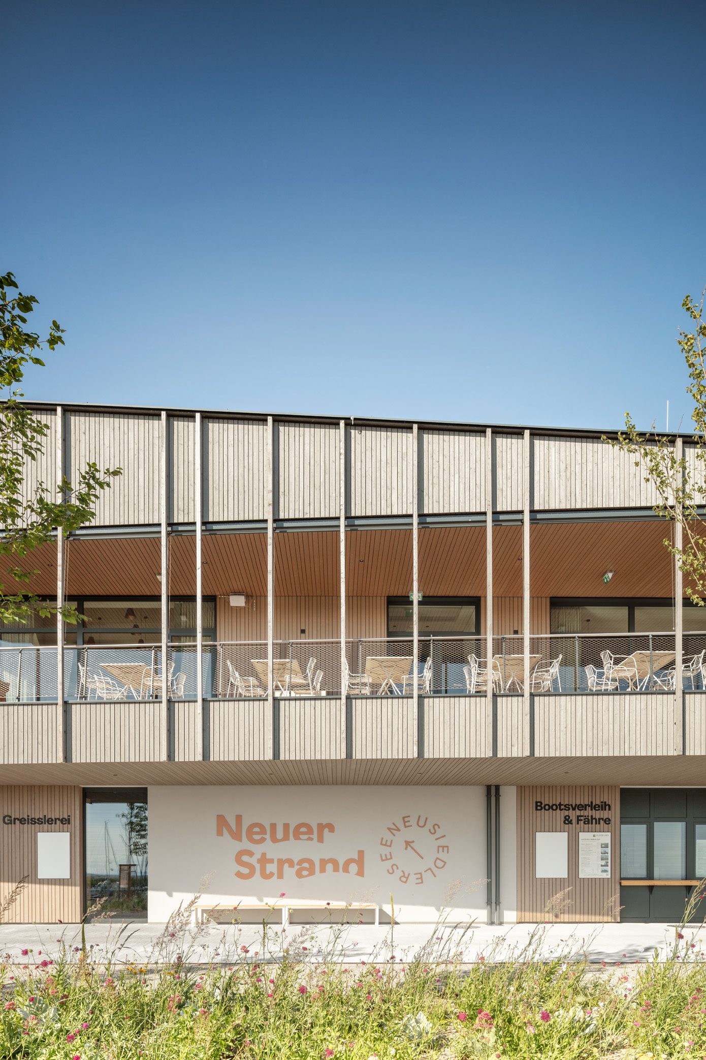 Modernes, zweigeschossiges Gebäude mit Holzlamellen-Fassade und großzügiger Terrasse im Obergeschoss. Erdgeschoss mit Schildern „Neuer Strand“ und „Bootsverleih & Fähre“. Minimalistische Architektur mit klaren Linien und naturnaher Gestaltung.