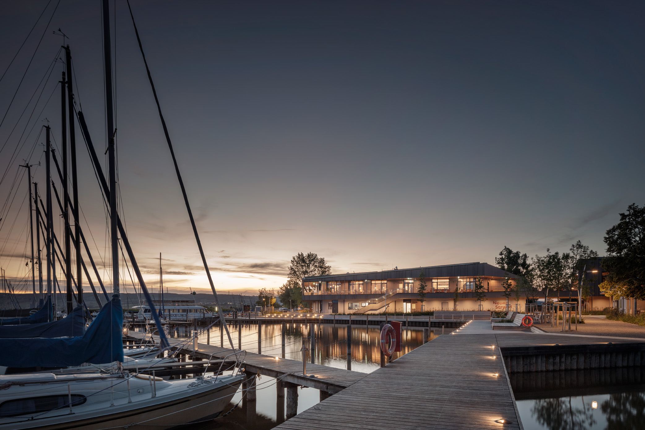 Moderner, zweigeschossiger Holzbau Neuer strand mit großen Glasflächen und vertikaler Fassadenstruktur, umgeben von einer naturnah gestalteten Gartenanlage mit Sitzbank und schattenspendendem Baum. Die Architektur kombiniert klare Linien mit nachhaltiger Begrünung. Abendstimmung mit Sundowner am Sonnendeck.