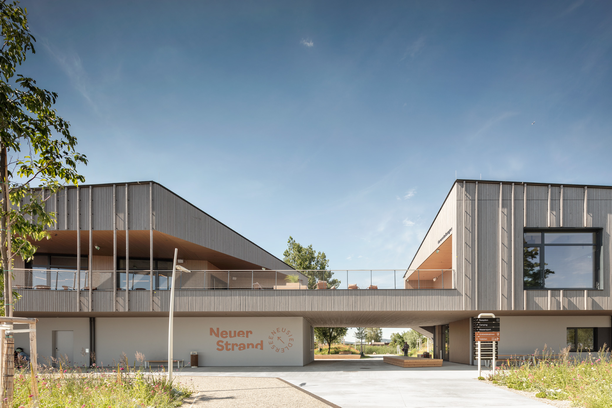 Modernes, zweigeschossiges Gebäude mit Holzfassade und großer Durchfahrt. Balkon mit Glasgeländer, große Fenster. Schriftzug „Neuer Strand“ an der Wand. Gepflasterter Weg, Infotafel und Pflanzen im Vordergrund. Blauer Himmel mit wenigen Wolken.