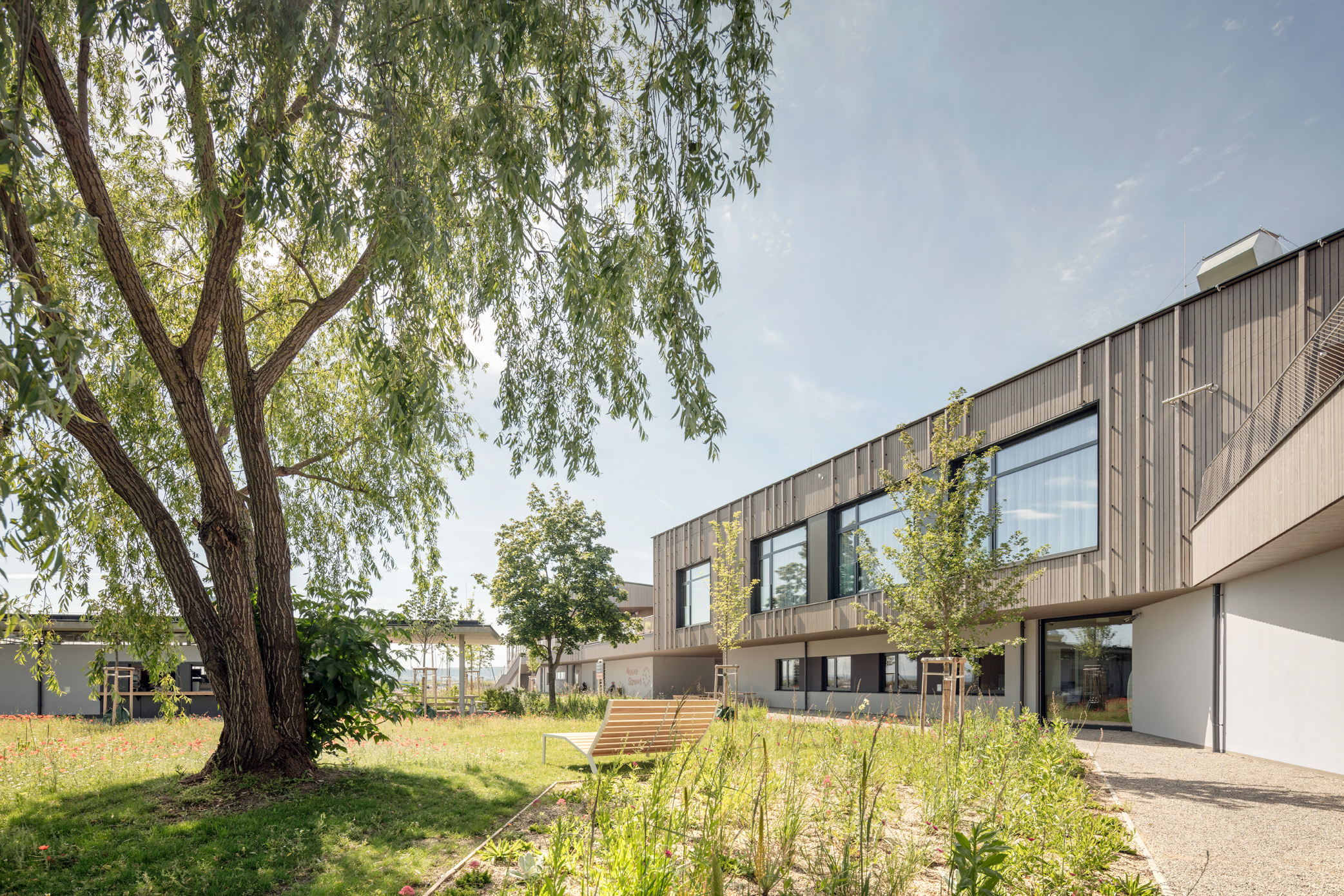 Moderner, zweigeschossiger Bau mit großen Glasflächen und vertikaler Fassadenstruktur, umgeben von einer naturnah gestalteten Gartenanlage mit Sitzbank und schattenspendendem Baum. Die Architektur kombiniert klare Linien mit nachhaltiger Begrünung.