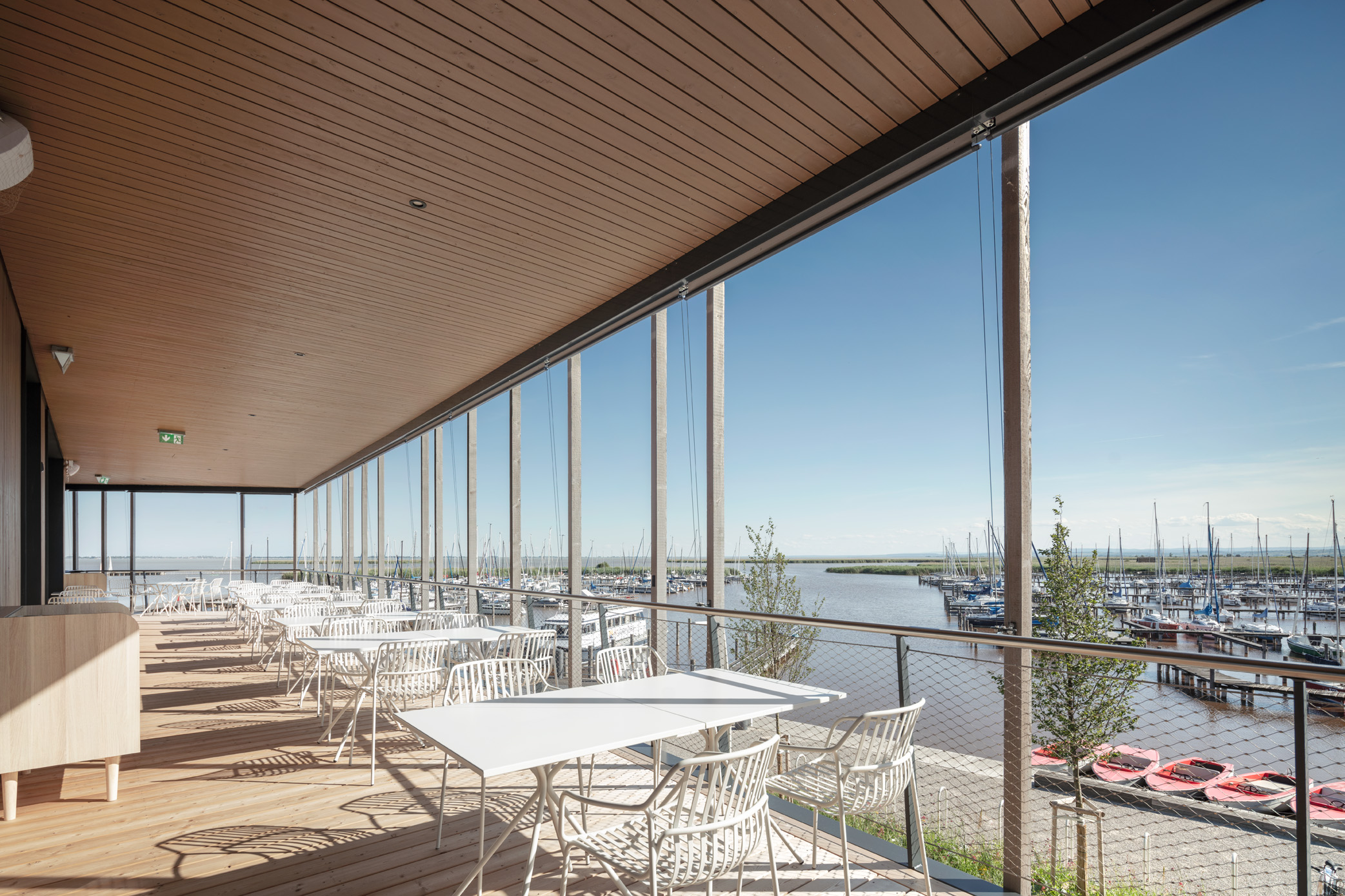 Moderne Seeterrasse des Restaurants Libelle mit weißen Outdoor-Möbeln, Holzverkleidung und Blick auf den Yachthafen – ideal für Gastronomie oder Veranstaltungen am Wasser.