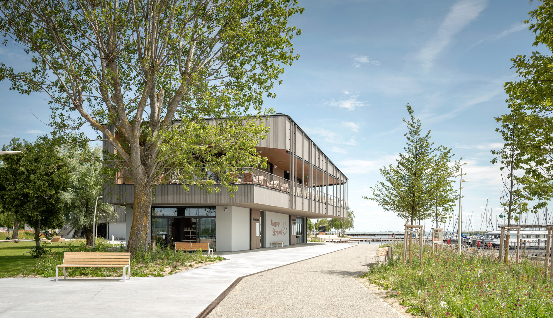 Modernes GebäudeNeuer Strand in naturnaher Umgebung mit Blick auf einen Yachthafen. Großzügige Terrasse im Obergeschoss, umgeben von Bäumen und Blumenwiese, lädt zum Verweilen ein. Breite Wege führen entlang der Wasserpromenade.