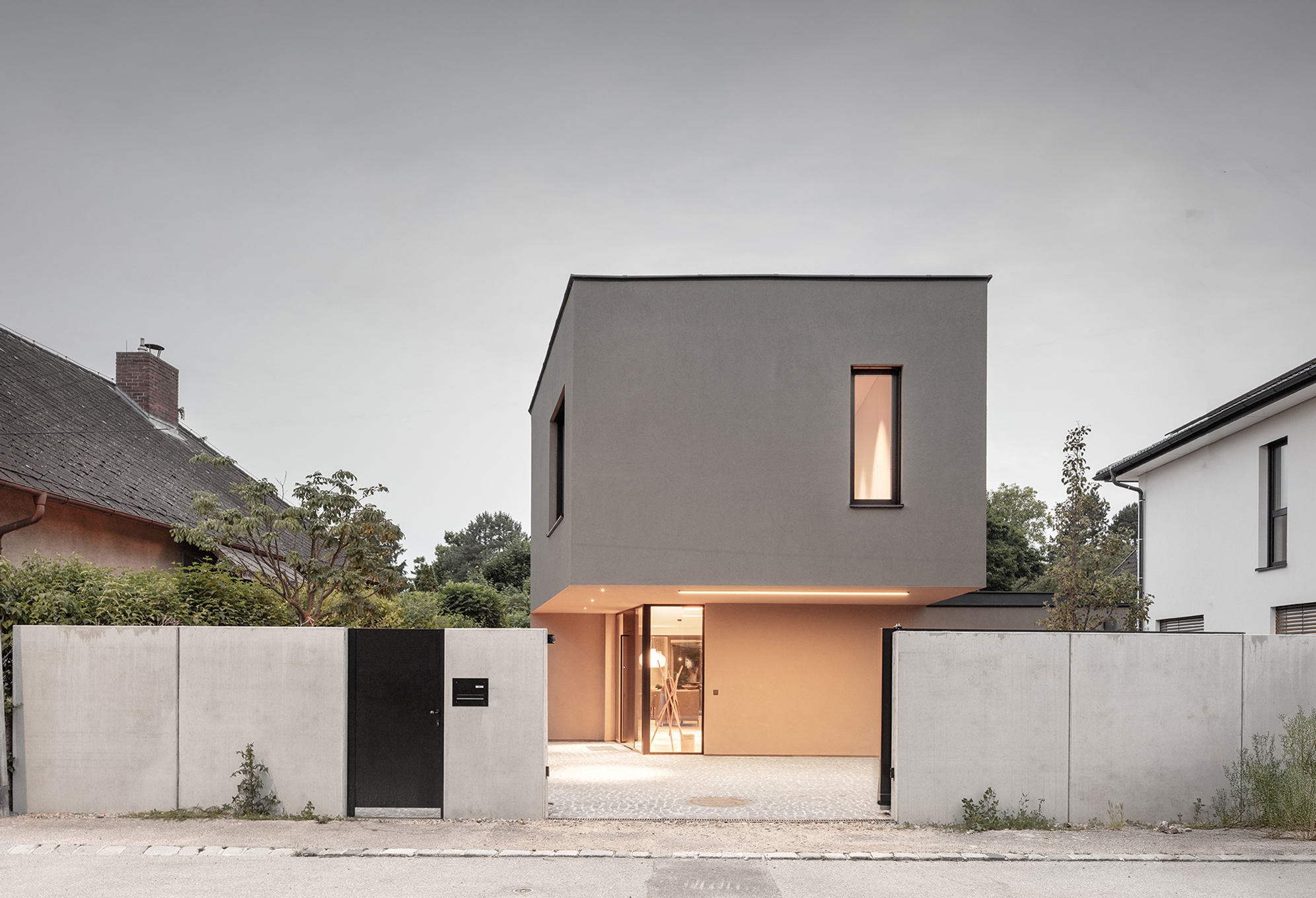 Kubisches Wohnhaus mit beige Putzfassade, schmalem Fenster und beleuchtetem Eingangsbereich zur Straße.