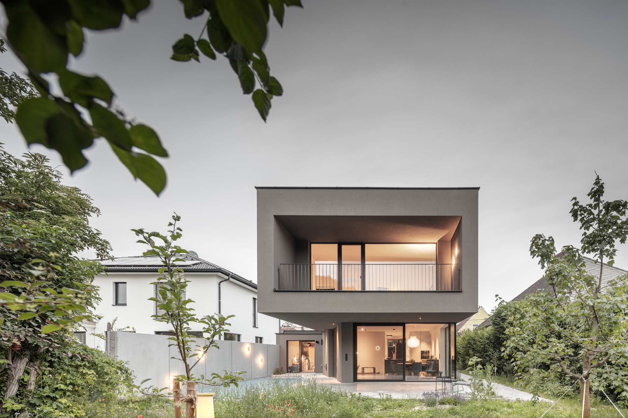 Modernes Wohnhaus mit auskragender Loggia, Glasfront und Garten mit Pool in der Dämmerung.